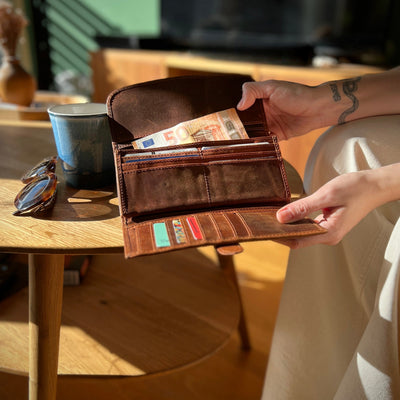 Person holding a brown leather wallet with money on a wooden table.