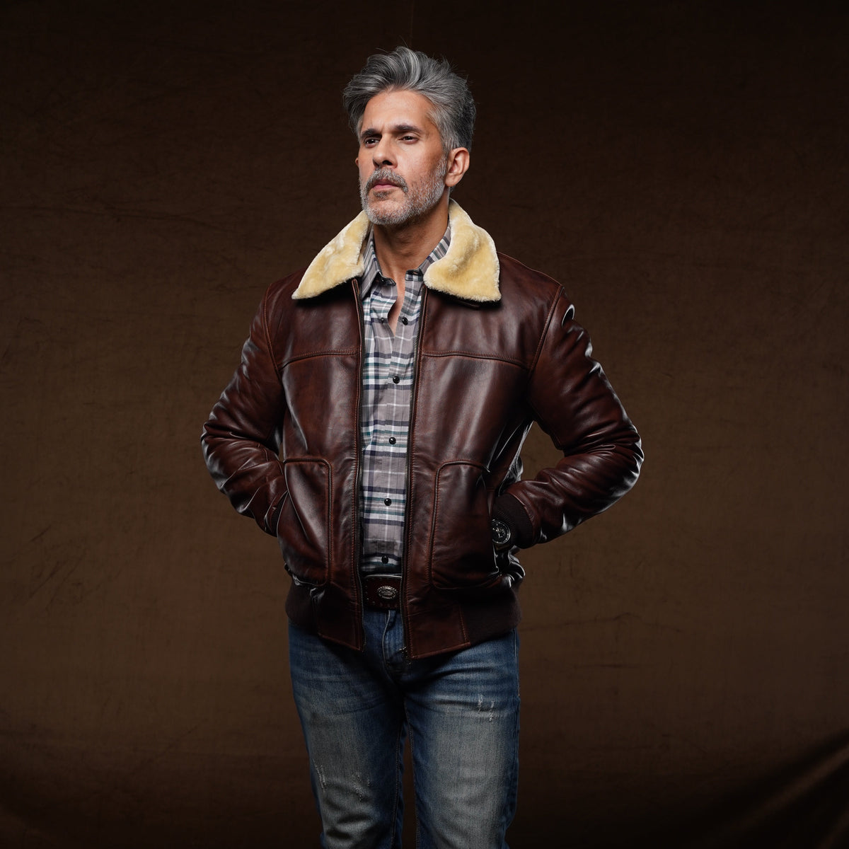 A man wearing a dark brown leather aviator-style jacket with a beige shearling collar, layered over a plaid shirt and blue jeans, standing against a dark brown studio backdrop.