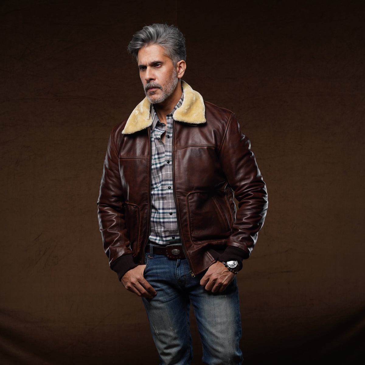 A man wearing a brown leather aviator-style jacket with a beige shearling collar, layered over a plaid shirt, blue jeans, and brown boots, standing against a dark brown studio backdrop.