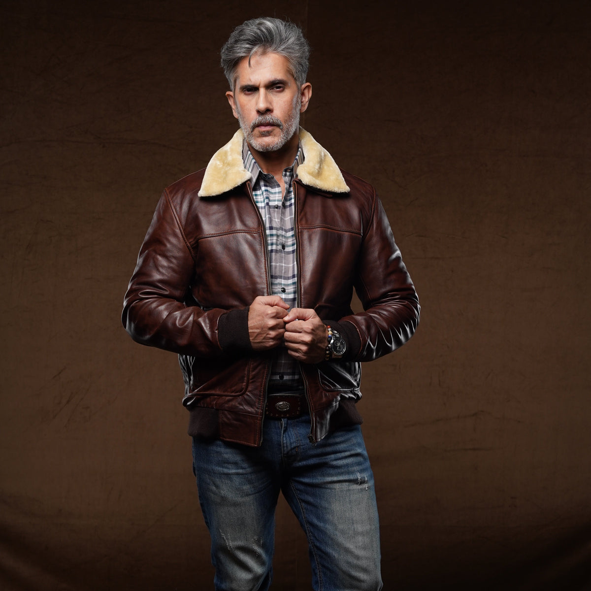 A man wearing a brown leather aviator-style jacket with a beige shearling collar, blue jeans, and brown boots, standing against a dark brown studio backdrop.