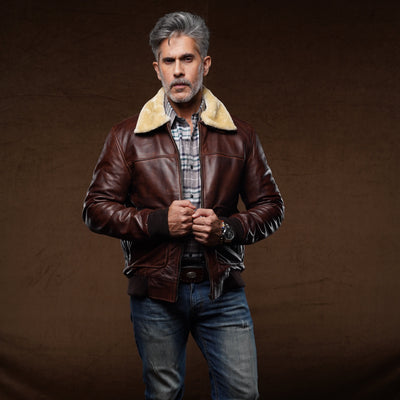 A man wearing a brown leather aviator-style jacket with a beige shearling collar, blue jeans, and brown boots, standing against a dark brown studio backdrop.