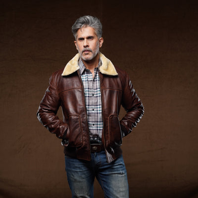 A man wearing a brown leather aviator-style jacket with a beige shearling collar, layered over a plaid shirt and blue jeans, standing against a dark brown studio backdrop.