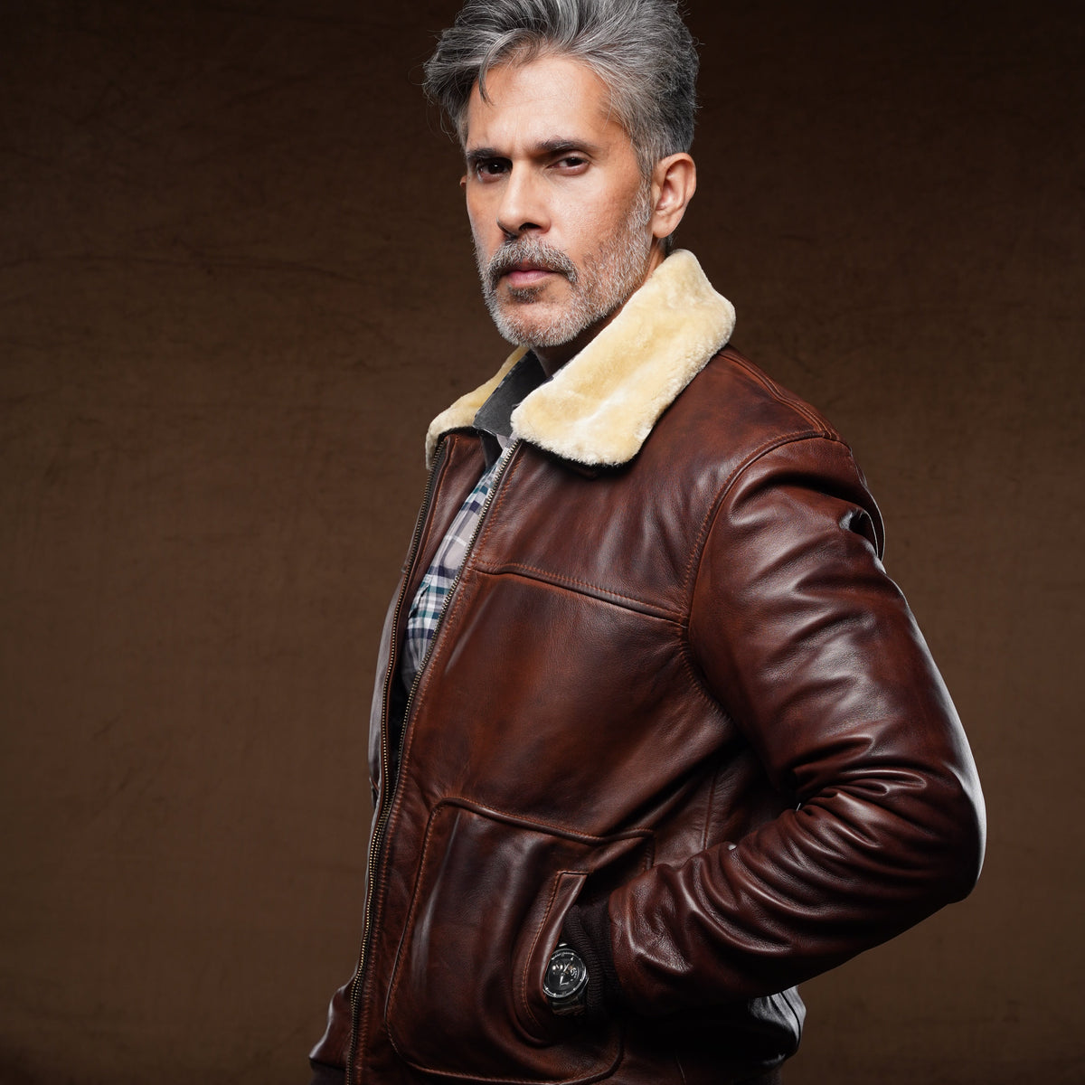 A man wearing a brown leather aviator-style jacket with a cream shearling collar, blue jeans, and a plaid shirt, posed against a dark brown studio background.
