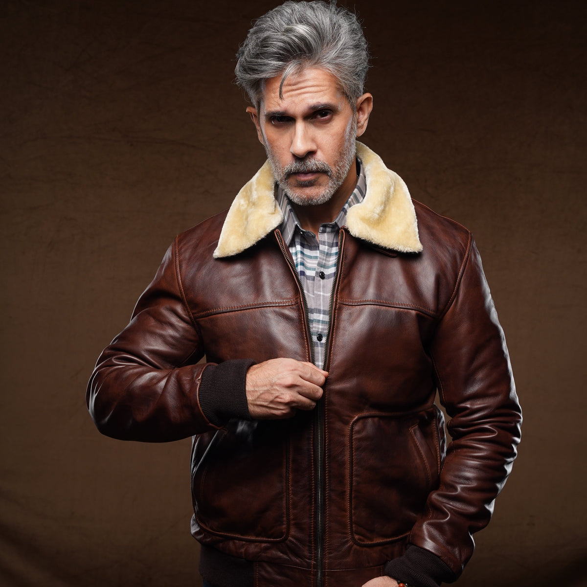 A man with gray hair and a beard wearing a brown leather aviator jacket with a beige shearling collar, blue jeans, and a plaid shirt, posing against a dark brown studio background.