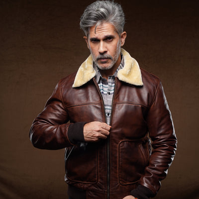 A man with gray hair and a beard wearing a brown leather aviator jacket with a beige shearling collar, blue jeans, and a plaid shirt, posing against a dark brown studio background.