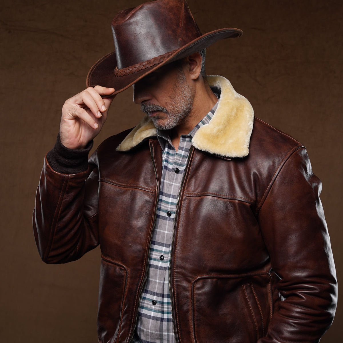 A man wearing a brown leather aviator-style jacket with a cream shearling collar, a brown cowboy hat, and a plaid shirt, posing against a dark brown textured studio background.