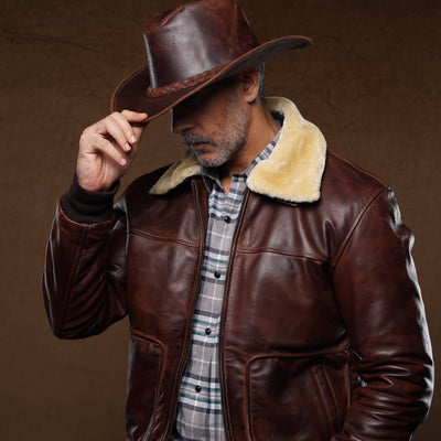 A man wearing a brown leather aviator-style jacket with a cream shearling collar, a brown cowboy hat, and a plaid shirt, posing against a dark brown textured studio background.
