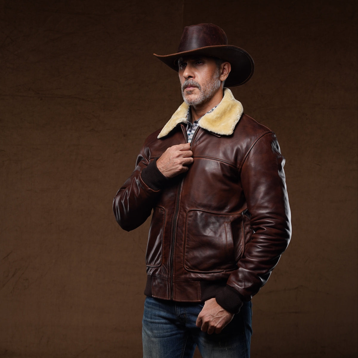 A man wearing a dark brown leather aviator jacket with a cream shearling collar, blue jeans, brown boots, and a brown cowboy hat, standing against a dark brown studio backdrop.