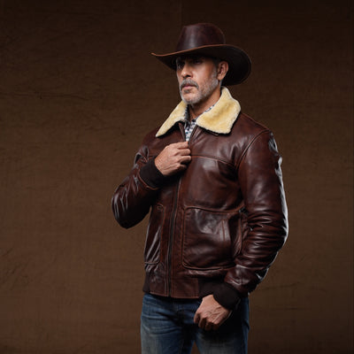 A man wearing a dark brown leather aviator jacket with a cream shearling collar, blue jeans, brown boots, and a brown cowboy hat, standing against a dark brown studio backdrop.