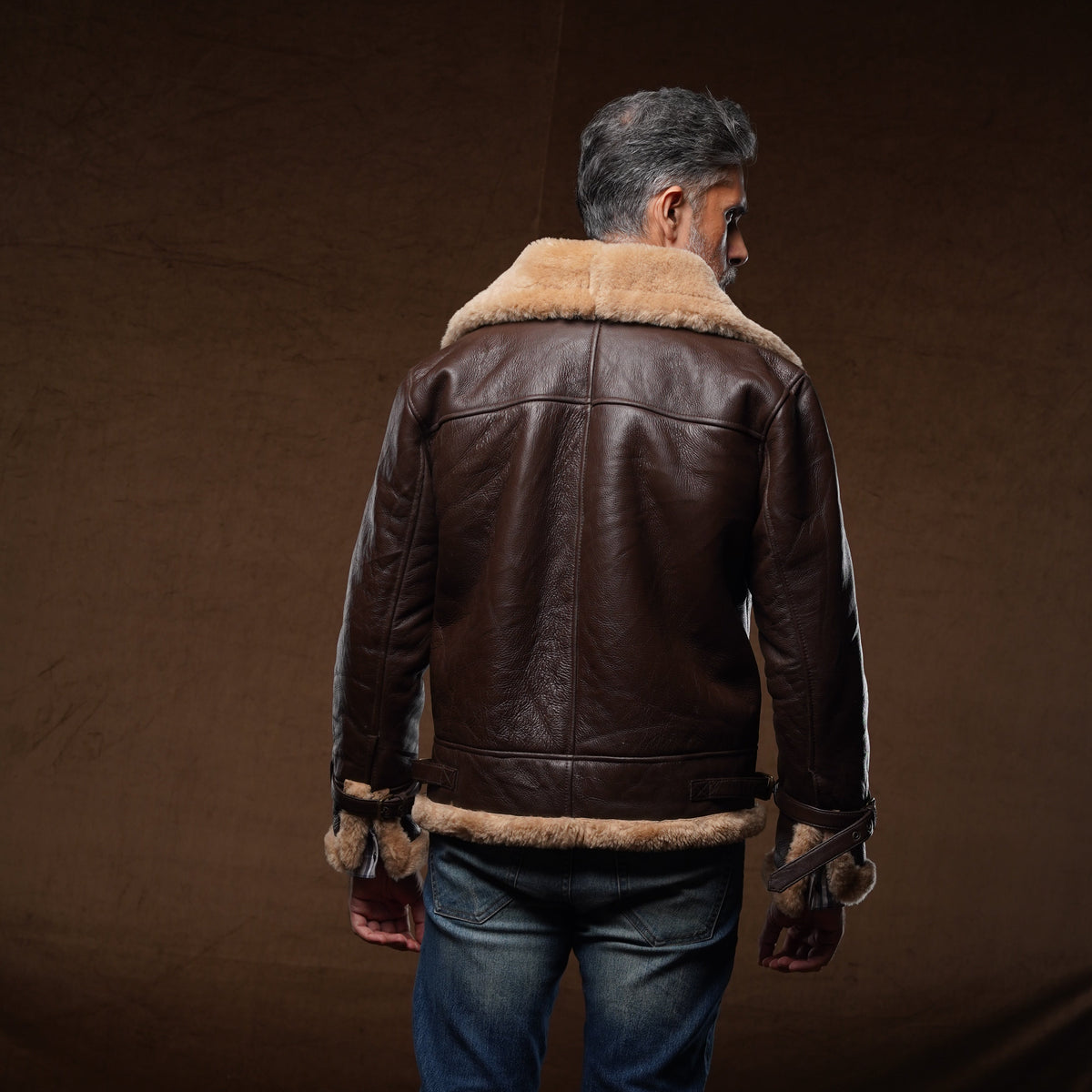 A man viewed from behind wearing a brown leather aviator jacket with a beige shearling collar, blue jeans, and brown boots, standing against a dark brown studio backdrop.
