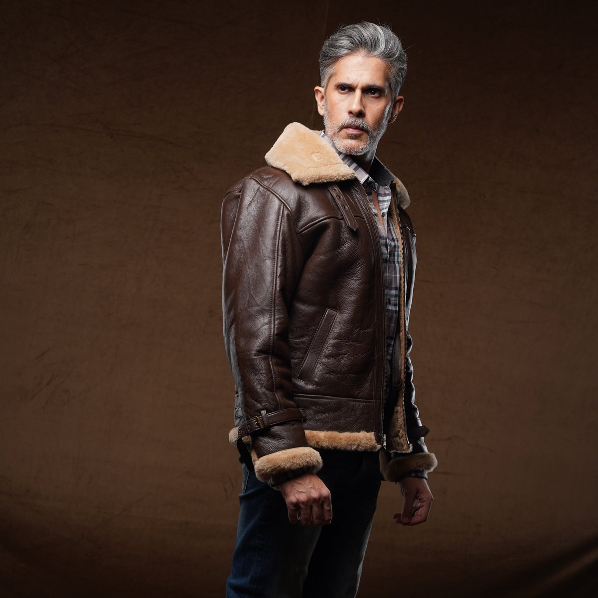 A man with gray hair and a beard wearing a brown leather aviator jacket with a beige shearling lining, blue jeans, and brown boots, standing against a dark brown studio backdrop.