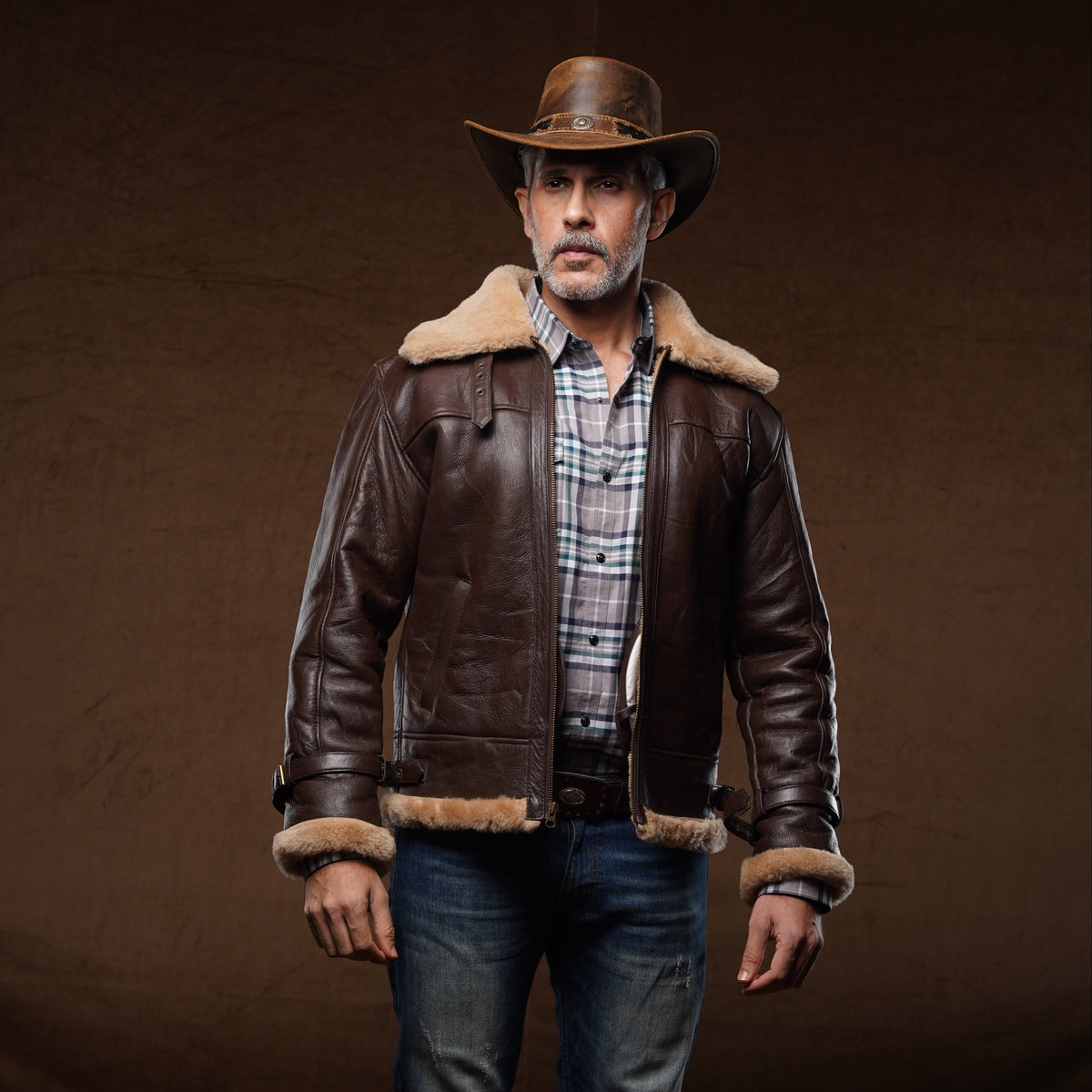 A man wearing a brown leather aviator-style jacket with a tan shearling collar, a brown cowboy hat, a plaid shirt, blue jeans, and brown boots, standing against a dark brown studio backdrop.