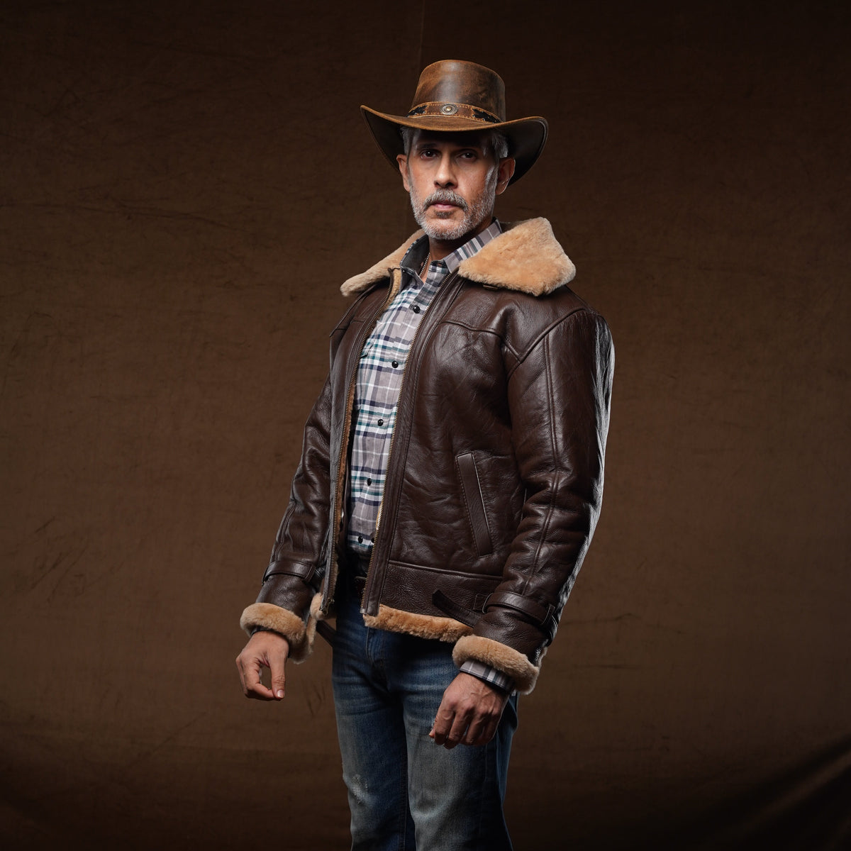 A man wearing a brown leather aviator-style jacket with a tan shearling collar, a brown cowboy hat, a plaid shirt, blue jeans, and brown boots, standing against a dark brown studio backdrop.