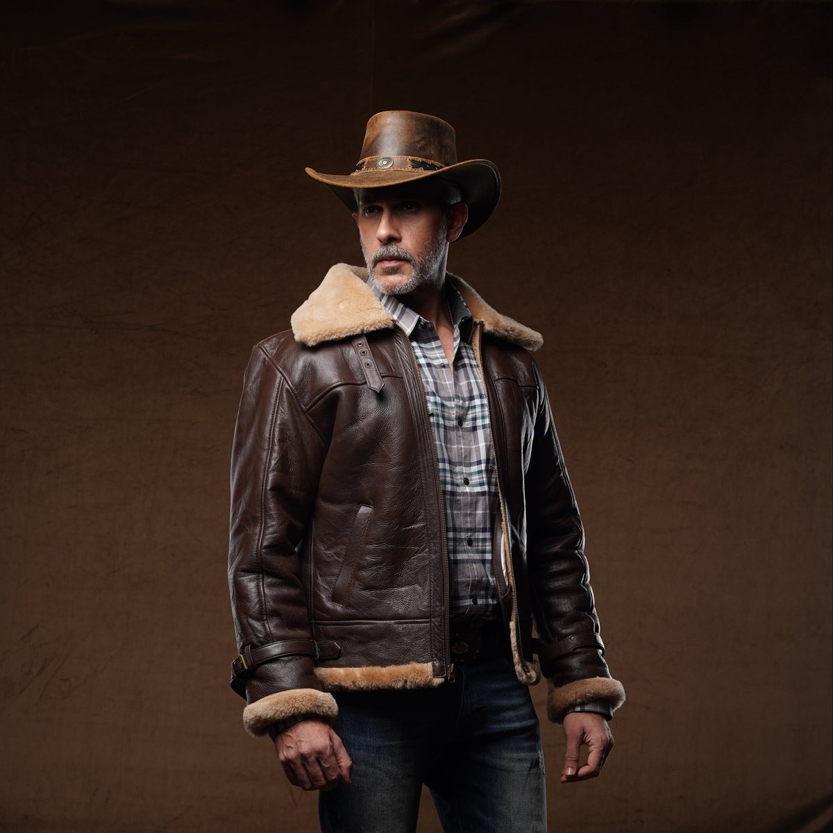 A man wearing a brown leather aviator-style jacket with a beige shearling collar, a brown cowboy hat, a plaid shirt, dark blue jeans, and brown boots, standing against a dark brown studio backdrop.